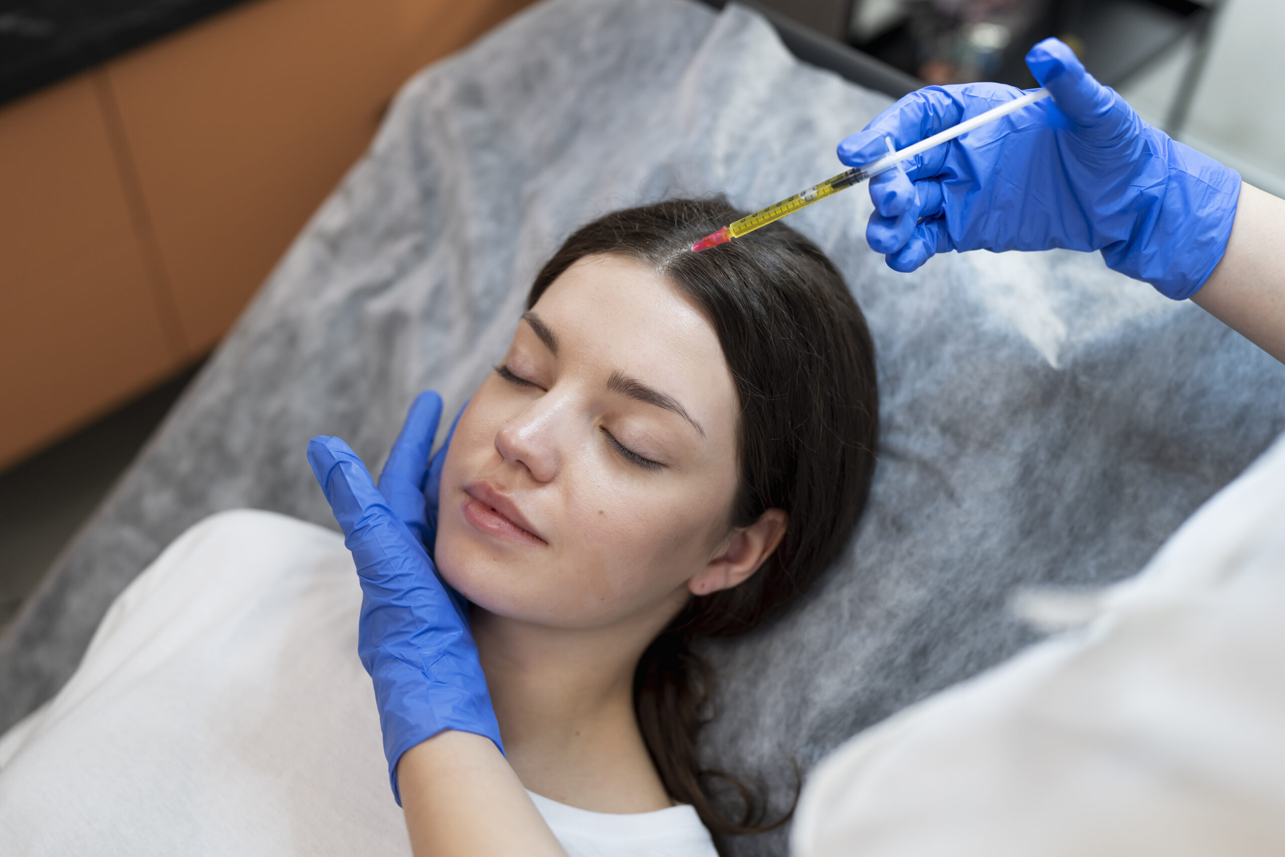 Medical professional in blue gloves administering a syringe injection to a woman's forehead as she rests on a clinic bed.
