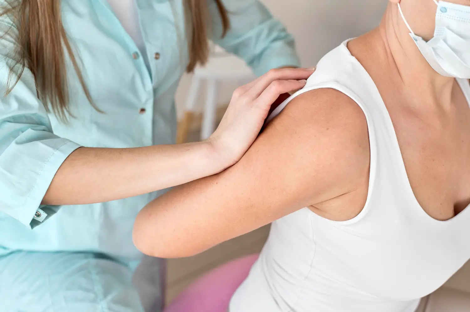 Medical professional in light blue scrubs rests a hand on a patient's shoulder during care.
